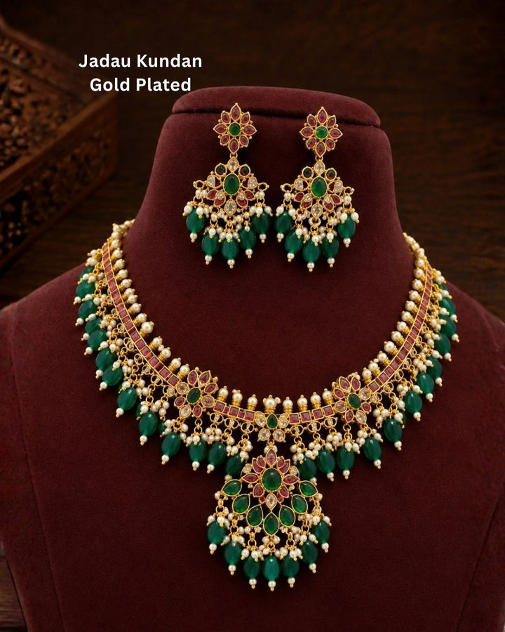 Traditional gold plated designer necklace and earrings set featuring intricate floral motifs with green and ruby-colored stones, sparkling white stones, pearl embellishments, and multiple green bead drops, perfect for festive, wedding, and traditional occasions.