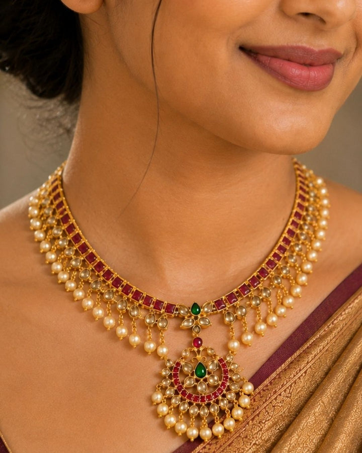 Beautiful Jadau Kundan gold plated jewellery set featuring a detailed necklace and matching earrings adorned with ruby, green stones, Kundan detailing, and pearl drops. Perfect for festive and traditional occasions.