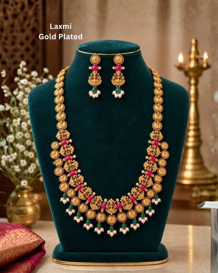 Elegant gold plated Lakshmi coin haram with detailed temple motifs, green beads, pearl drops, and matching Lakshmi earrings displayed on a teal jewellery bust.