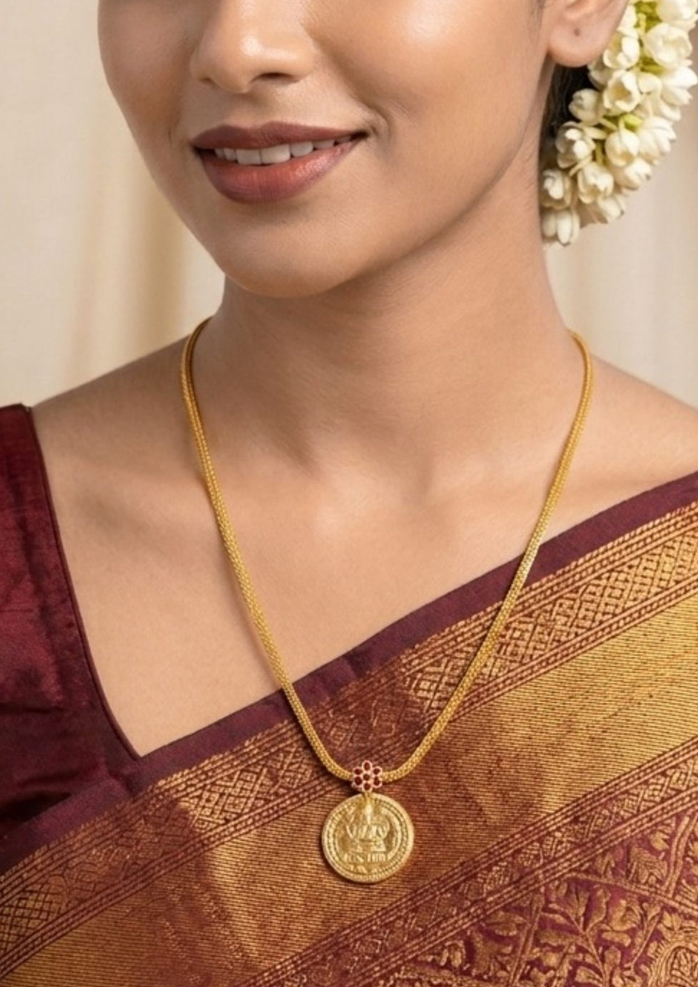 Woman wearing a gold necklace with a coin pendant Laxmi chain gold plated traditional necklace featuring a circular coin pendant embossed with Goddess Lakshmi seated on a lotus, connected by a small pink ruby stone floral accent to a thick textured mesh chain. This classic South Indian style necklace is crafted with a premium gold finish that provides a rich divine appearance suitable for weddings, festivals, poojas, family functions, Varalakshmi Vratam, Diwali, sarees, and ethnic outfits with skin-friendly