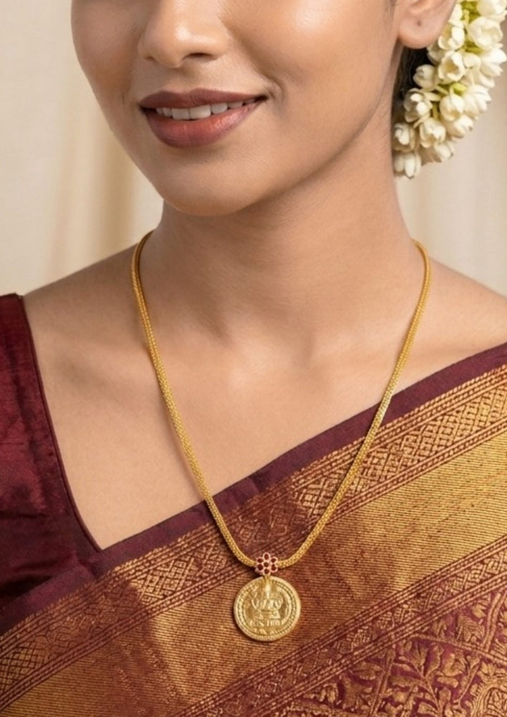 Woman wearing a gold necklace with a coin pendant Laxmi chain gold plated traditional necklace featuring a circular coin pendant embossed with Goddess Lakshmi seated on a lotus, connected by a small pink ruby stone floral accent to a thick textured mesh chain. This classic South Indian style necklace is crafted with a premium gold finish that provides a rich divine appearance suitable for weddings, festivals, poojas, family functions, Varalakshmi Vratam, Diwali, sarees, and ethnic outfits with skin-friendly