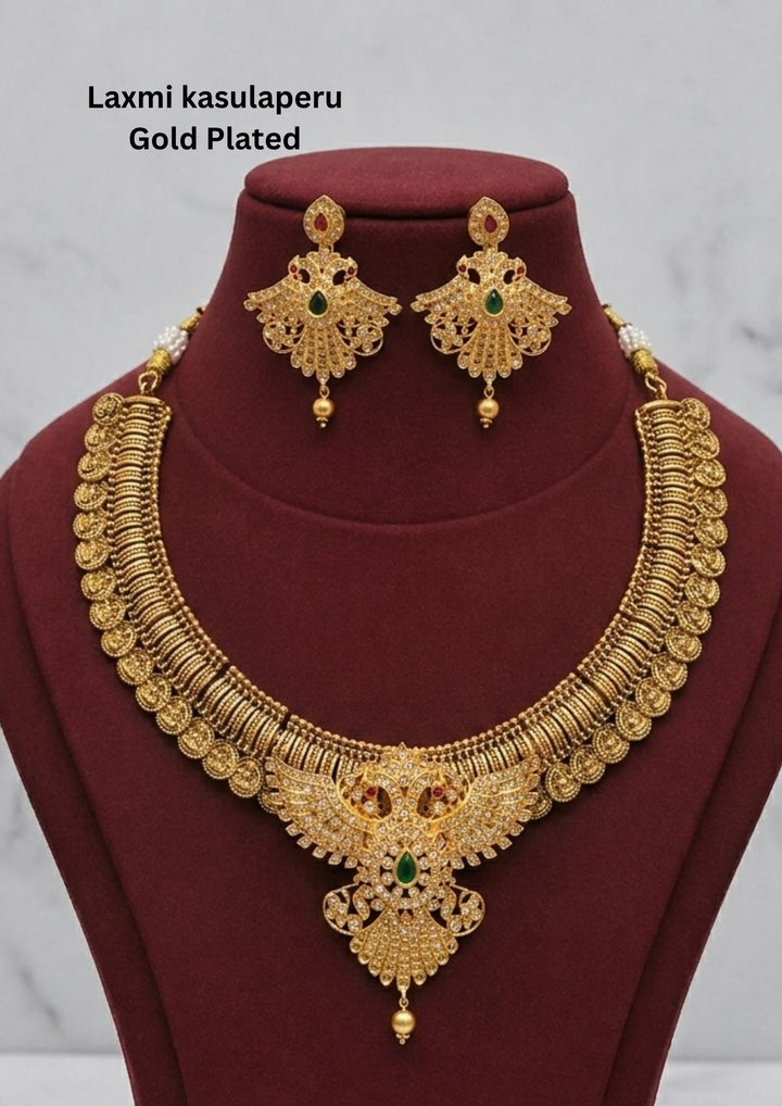 Traditional Laxmi kasulaperu gold plated short haram necklace featuring detailed coin style motifs throughout the chain with an intricately designed center pendant studded with white stones and green stone accents. The matching earrings have elegant filigree detailing with hanging gold beads, creating a rich South Indian temple jewelry appearance suitable for weddings, poojas and bridal occasions.