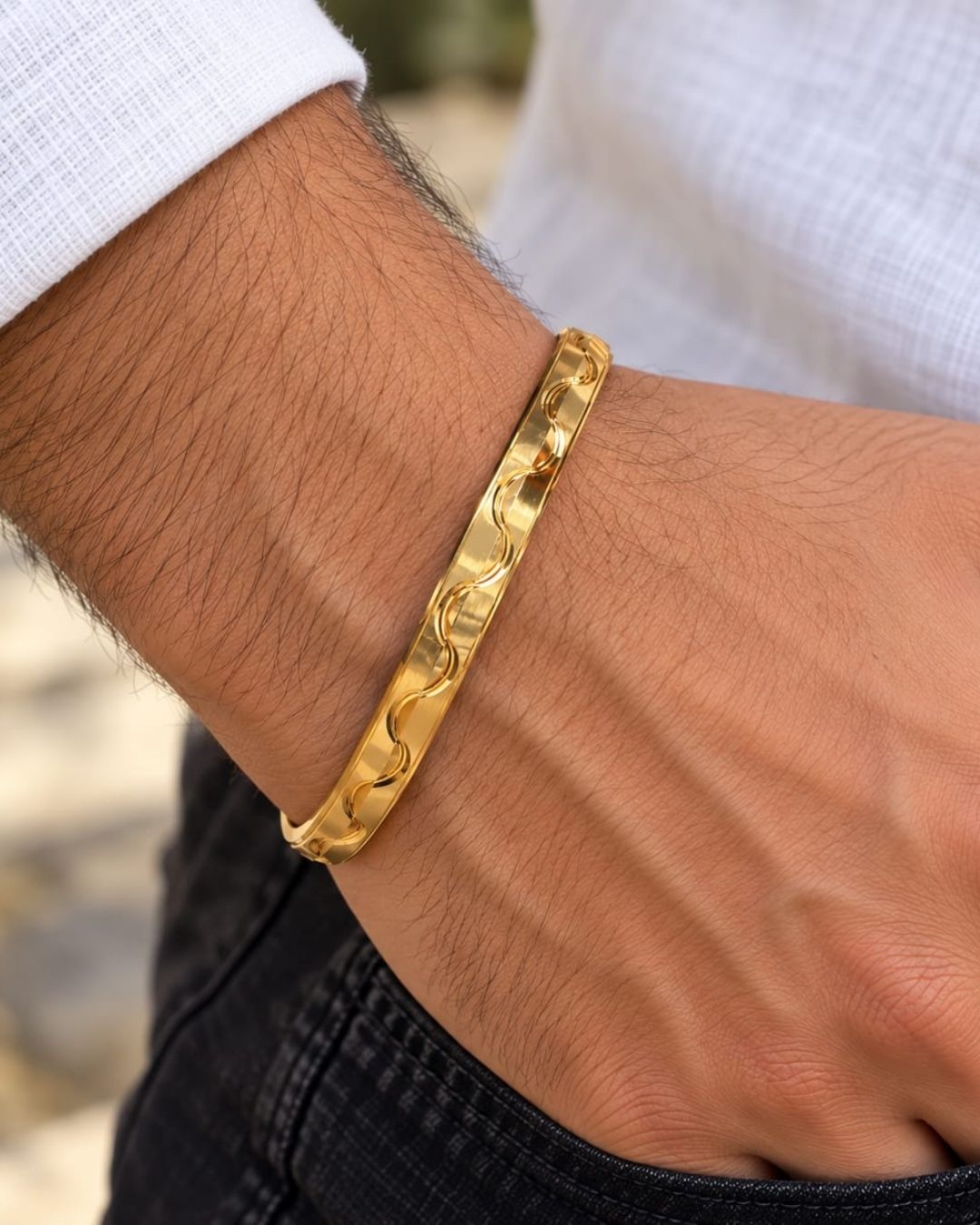Gold plated men’s kada displayed upright on a beige surface, showcasing its modern wave pattern and reflective finish.
