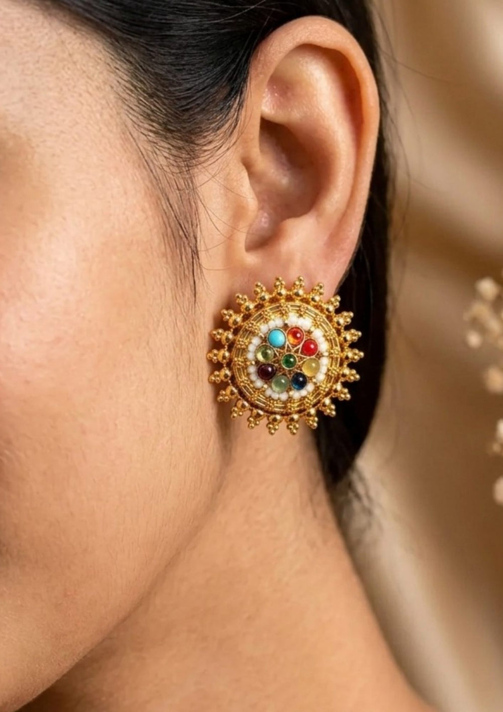 Close-up of a gold earring with colorful stones gold plated navaratna style round stud earrings featuring a wheel pattern arrangement of vibrant multi-color stones including turquoise blue, ruby red, emerald green, amethyst purple, and amber yellow bordered by fine white beads and an outer sunburst gold bead trim. These lightweight large statement studs provide a grand traditional look suitable for weddings, festivals, poojas, cultural events, festive sarees, lehengas, and ethnic outfits with skin-friendly 