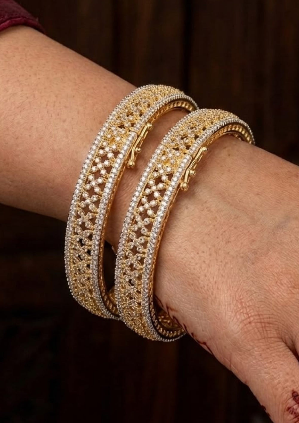Gold plated zircon diamond bangles on a wrist women wearing a stunning pair of 18k gold plated bangles featuring intricate open lattice craftsmanship with rows of shimmering white zircon stones along the borders. The broad kada-style bangles showcase a luxurious diamond-like finish with secure clasp closures, making them ideal for weddings, festive occasions, and traditional outfits. 