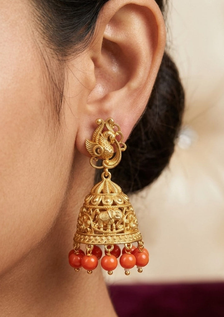 Gold earrings with red beads worn by a person Traditional coral jhumkas gold plated earrings featuring intricate temple-inspired craftsmanship with beautifully carved elephant motifs on the dome-shaped jhumka. The earrings are adorned with vibrant coral red bead danglers and detailed antique gold finish patterns, creating a rich South Indian ethnic look suitable for weddings, festivals, poojas and cultural celebrations.