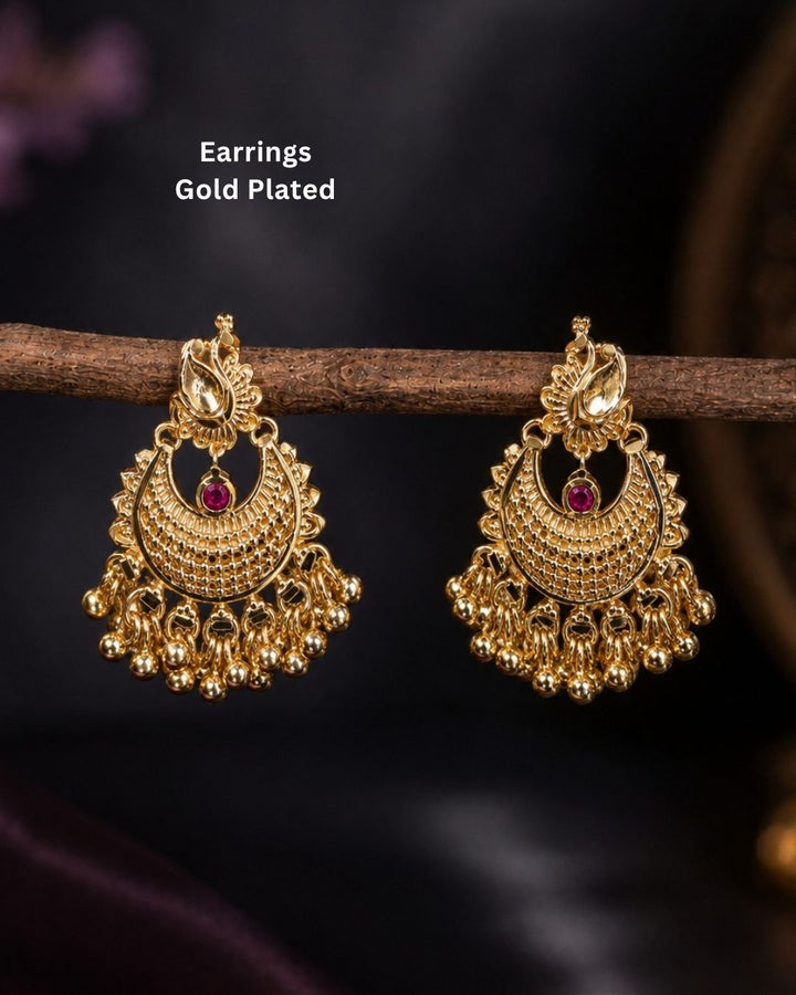 A pair of gold-plated traditional Indian jhumka earrings with intricate filigree detailing and a small red stone accent, hanging from a wooden branch against a dark background. White text in the upper left reads &