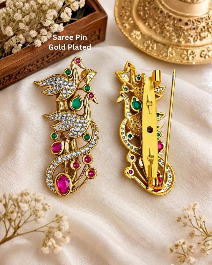 Pair of gold plated double peacock saree pins displayed on a light wooden surface, showcasing sparkling stones and decorative pin-back fastenings.
