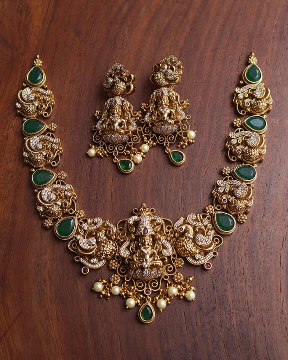 Premium antique gold plated Lakshmi necklace and earrings set crafted with intricate temple-inspired detailing, sparkling stones, green accents, and pearl embellishments.