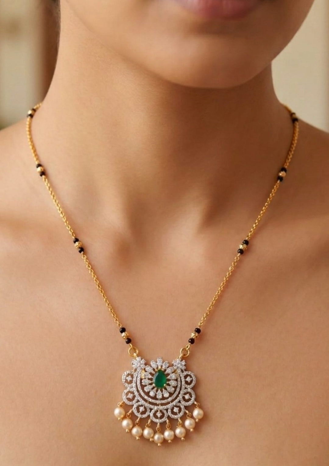 Gold necklace with a green gemstone pendant Modern Zircon Diamond gold plated mangalsutra necklace The design features a delicate gold link chain transitioning into sections of traditional small black beads on both sides. The central pendant showcases a sleek, contemporary curved u-shape and a stylized butterfly or floral motif encrusted with sparkling white cubic zirconia stones. This elegant neckpiece perfect for daily wear, office wear, and casual outfits.