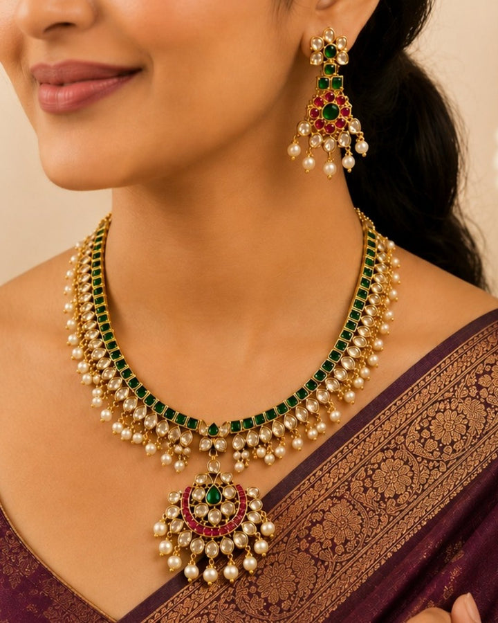 A south indian woman wearing an ornate gold-toned traditional jewelry set with deep red stones, including a necklace, matching earrings, and a forehead ornament, paired with a navy blue embroidered saree.