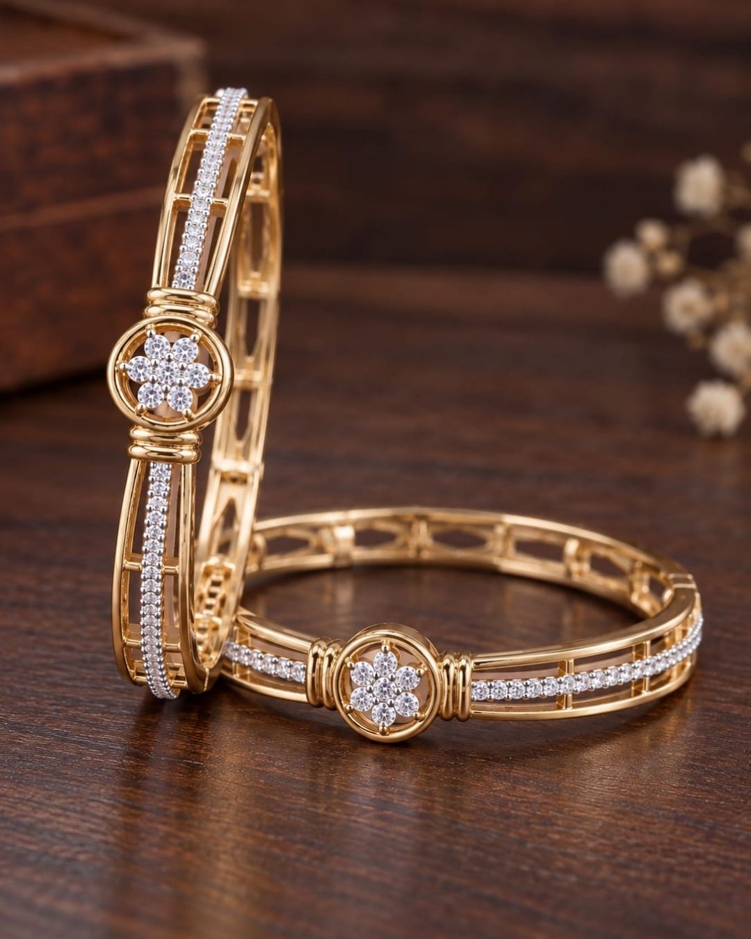 Pair of gold plated designer bangles displayed upright on a dark wooden surface with a rich brown background.