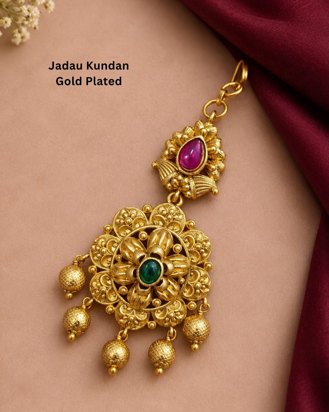 Traditional gold plated designer maangtikka featuring intricate floral motifs, green and ruby-colored stones, detailed gold work, and multiple hanging gold bead accents, perfect for festive, wedding, and traditional occasions.