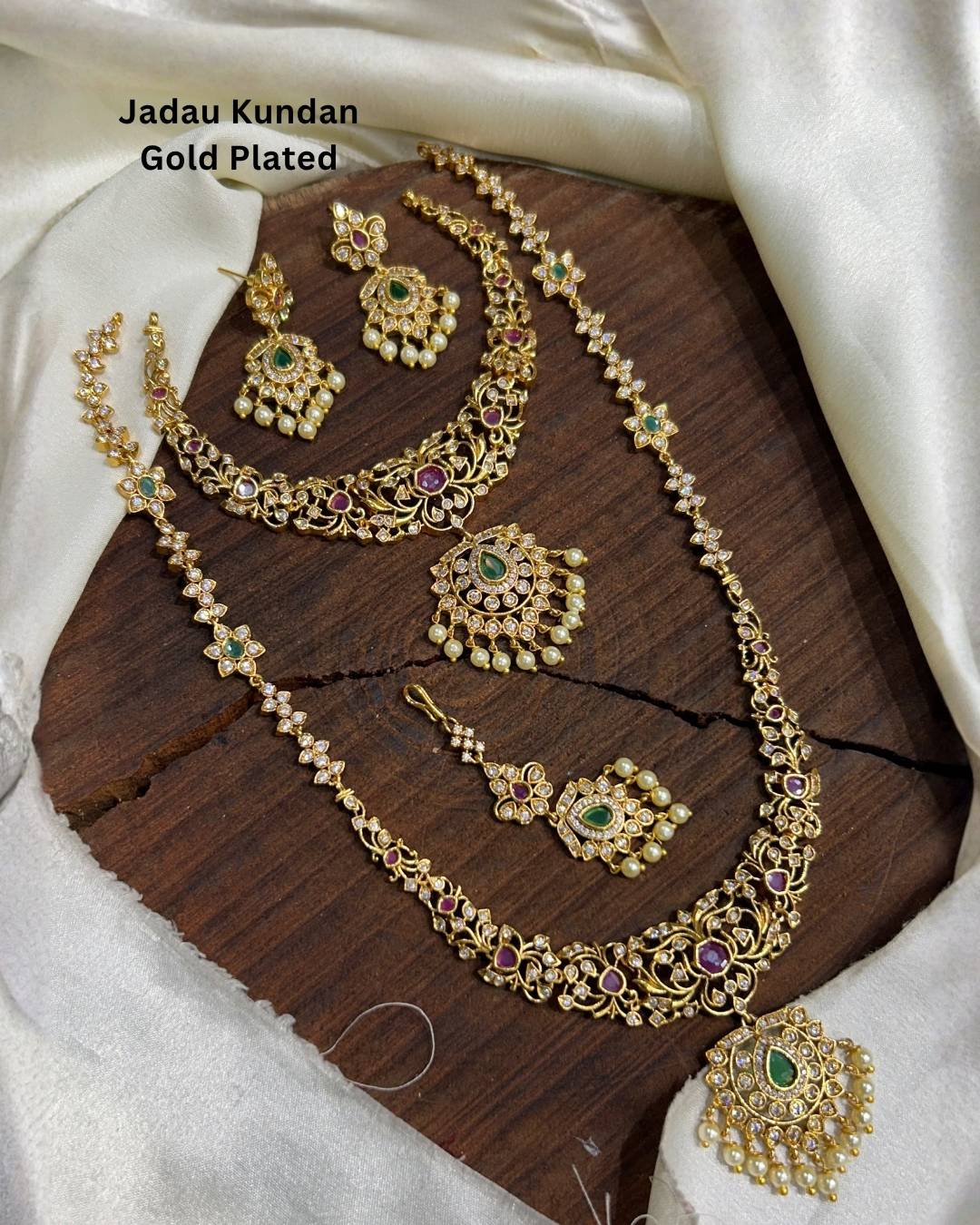 Traditional gold plated wedding jewelry collection including layered necklaces, matching earrings, maang tikka, and pendant with green stones, ruby accents, and pearl embellishments.