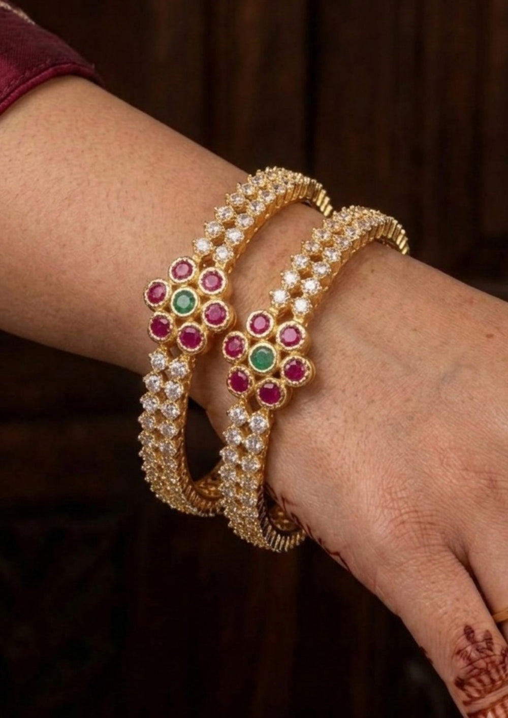 Gold bangles with gemstones on a wrist  Zircon diamond gold plated bangles pair featuring multiple rows of glistening white zircon stones highlighted by a prominent central floral cluster made of rich ruby pink petals and a green emerald core. This premium kada style set provides a heavy statement look while remaining lightweight and comfortable, suitable for weddings, festivals, receptions, family gatherings, sarees, lehengas, and ethnic outfits with skin-friendly quality, anti-fade polish, secure fitting,