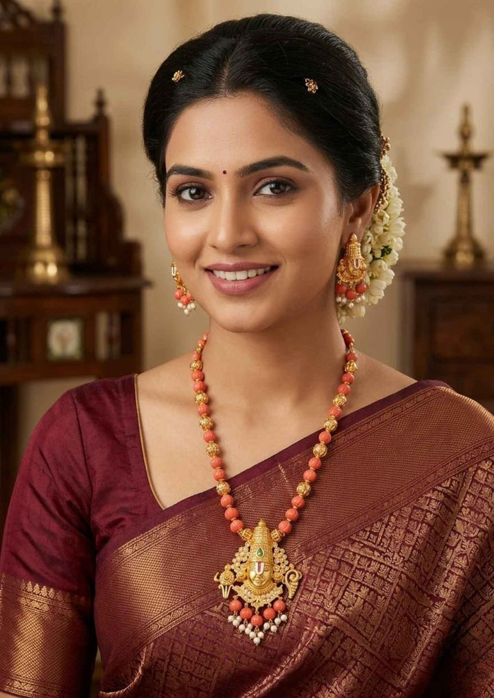 A South Indian woman wearing a Gold Plated Balaji Coral Beads necklace set with matching earring set with a polished gold finish, The design highlights a beautifully carved blalaji pendant suspended from a neat coral bead strand, The long coral mala and matching earrings pattern with Lord Venkateswara designs, pearl drops, guttapusalu and a smooth finish, Perfecr for festivals, poojas, and weddings