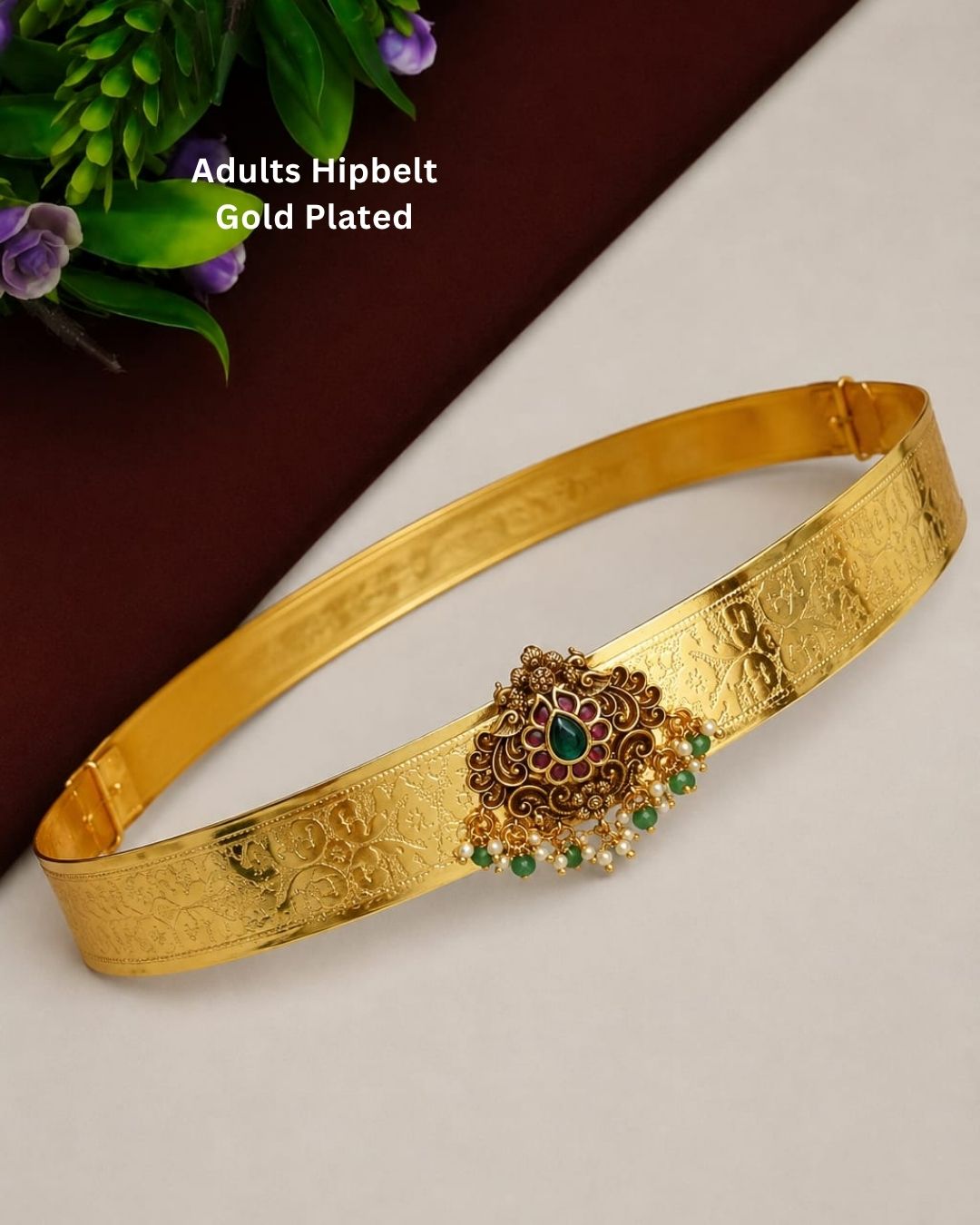 Beautiful gold plated adult hip belt featuring a wide intricately engraved band with a floral gemstone centerpiece decorated with ruby, green stones, and pearl drops. Perfect for traditional wear, weddings, festive occasions, bridal styling, parties, and gifting.