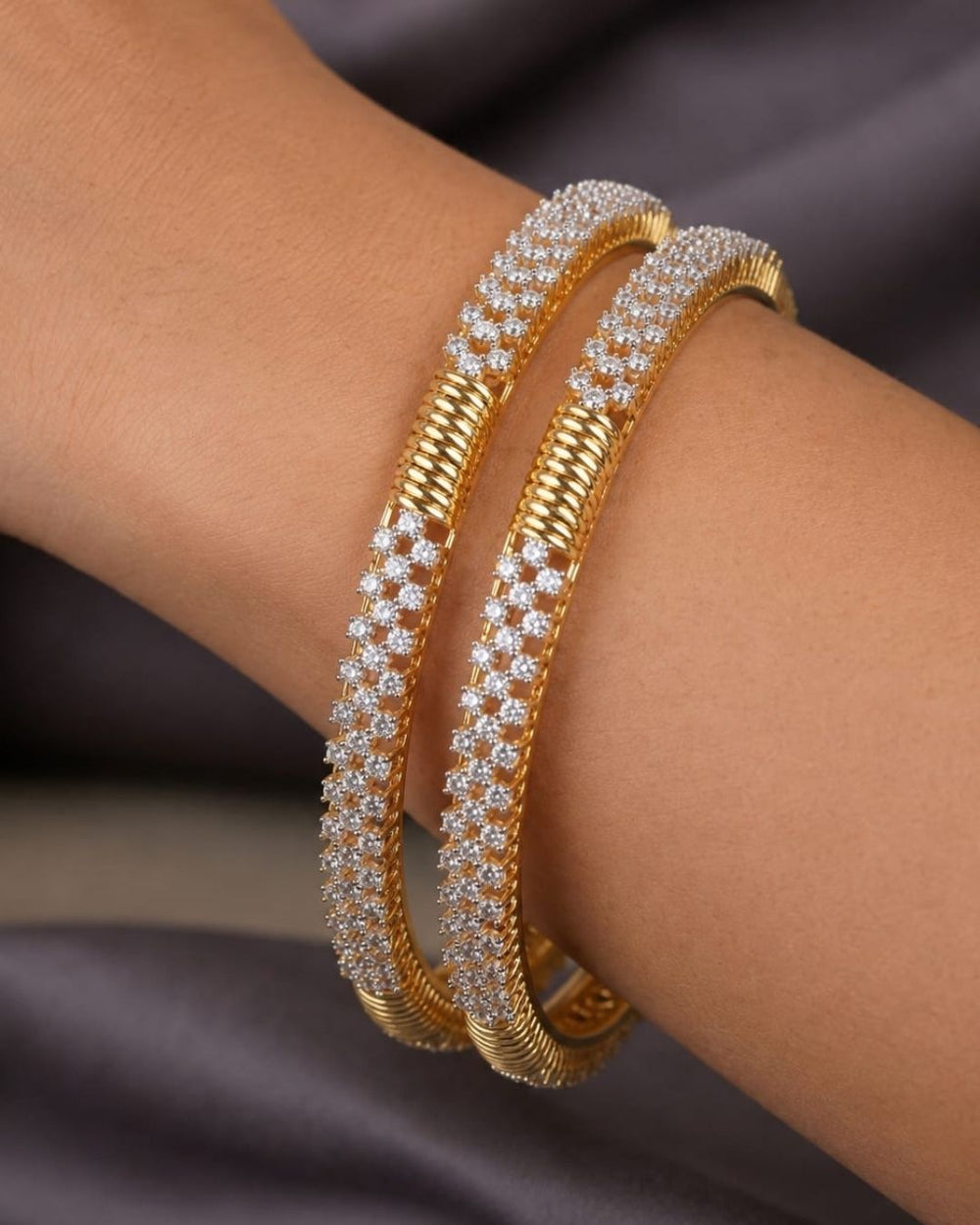 A pair of gold-plated huggie bangles adorned with rows of sparkling clear zirconia stones, displayed on a neutral surface with a blurred wooden background. Text overlay reads 'Zircon Diamond Gold Plated'.