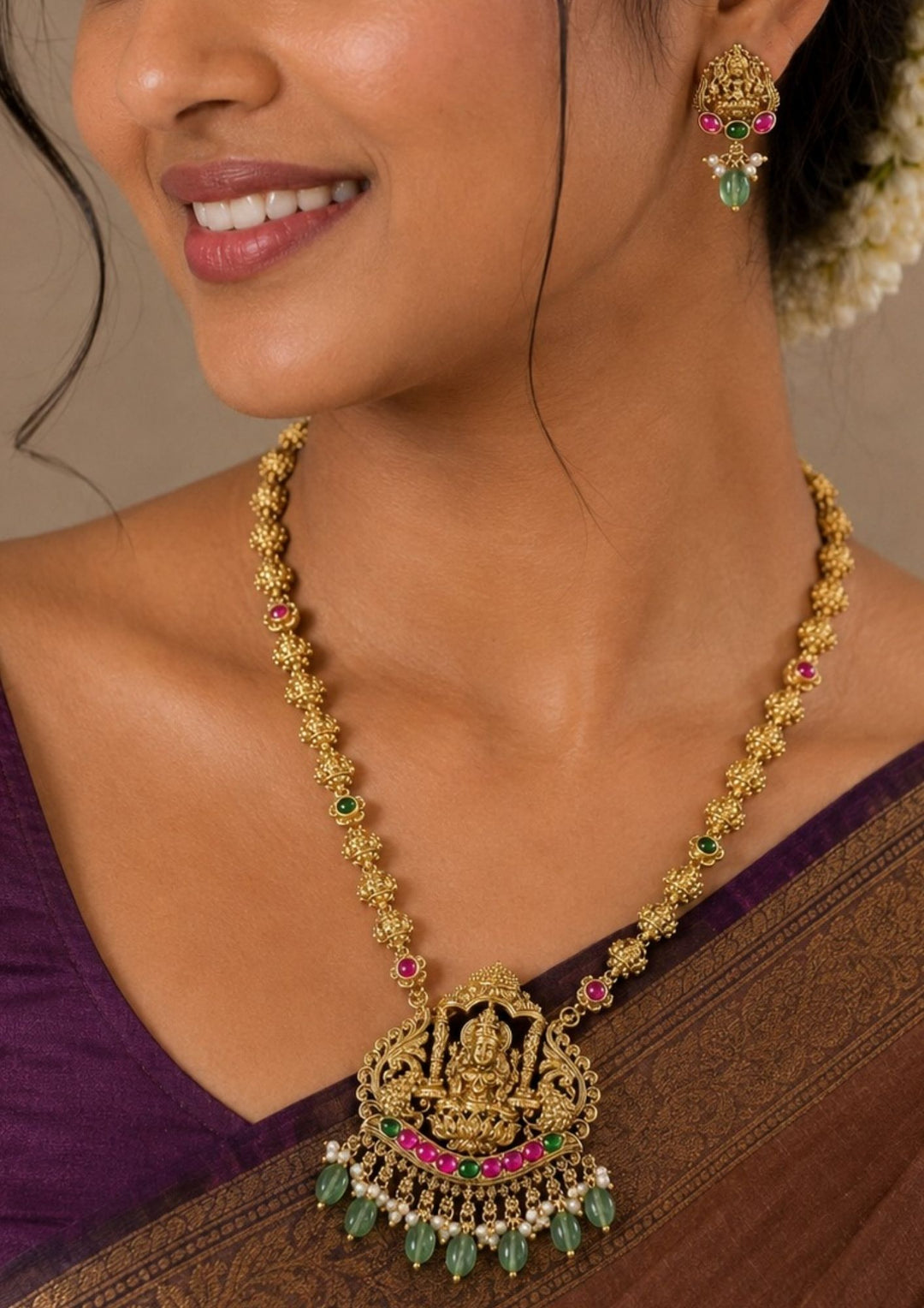 South Indian Woman wearing a Gold-plated laxmi devi necklace set with intricate design long necklace Set and earrings with green and red stones come with jadau kundan in necklace set  with beads and in laxmi devi pendent and earrings shows clear design and temple jewelry in detailed shown in picture it will not fade color and water proof can used for festivals, bridals, and weeding&