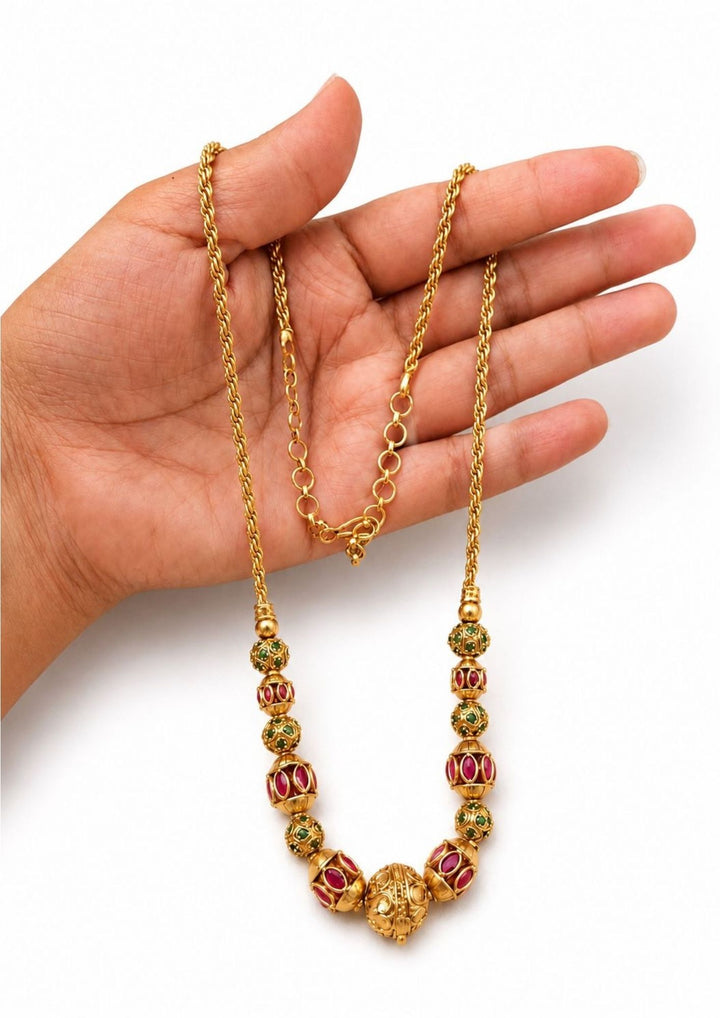 Gold plated long chain haraam necklace featuring elegant textured golden spheres alternating with small spacer rings inset with red ruby and green emerald stone accents. This lightweight chain is crafted with a detailed twisted rope style chain extension held in hand to provide a classy and premium appearance suitable for daily wear, office wear, casual outings, family gatherings, festivals, parties and traditional occasions with skin friendly quality, anti fade polish, comfortable fitting