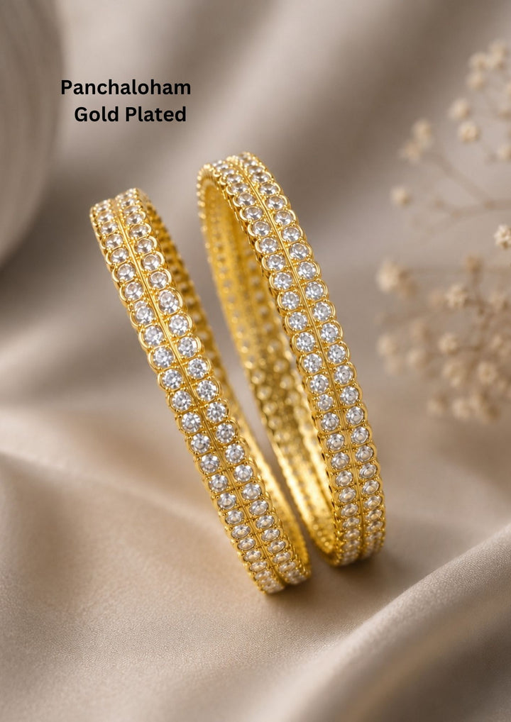 A pair of premium gold-plated bangles meticulously studded with two parallel rows of brilliant, round-cut clear zircon stones. Each crystal is secured in a classic prong setting along the outer rim of the band, creating a continuous, shimmering diamond-like effect against the rich, polished gold finish suitable for weddings, poojas and bridal occasions.