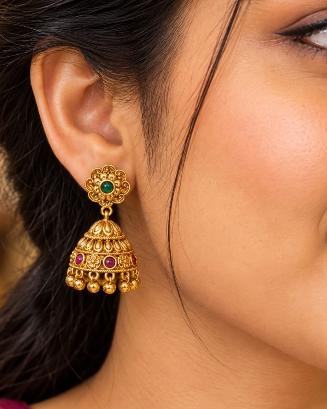 Traditional gold plated jhumka earrings featuring intricate floral motifs, green center stones, ruby-colored embellishments, detailed dome-shaped drops, and hanging gold beads, perfect for festive and traditional occasions.