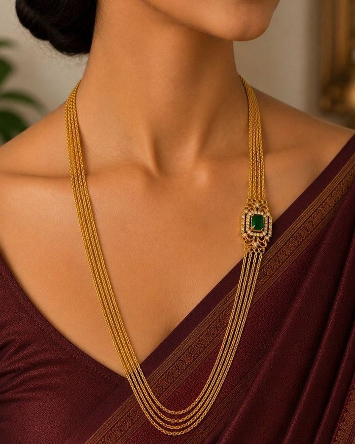 Beautiful green stone Chandra Haram gold plated necklace featuring multiple layered chains and an elegant rectangular green centerpiece with intricate detailing and sparkling stones. Perfect for festive occasions, traditional outfits, weddings, temple visits, and premium gifting.