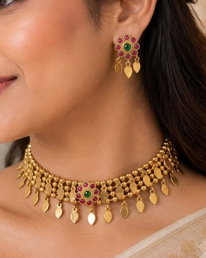 Gold-toned traditional Indian jewelry set featuring a choker necklace and matching drop earrings worn by a model. The jewelry is adorned with gold leaf-shaped charms, small beads, and central floral motifs set with green and red stones.