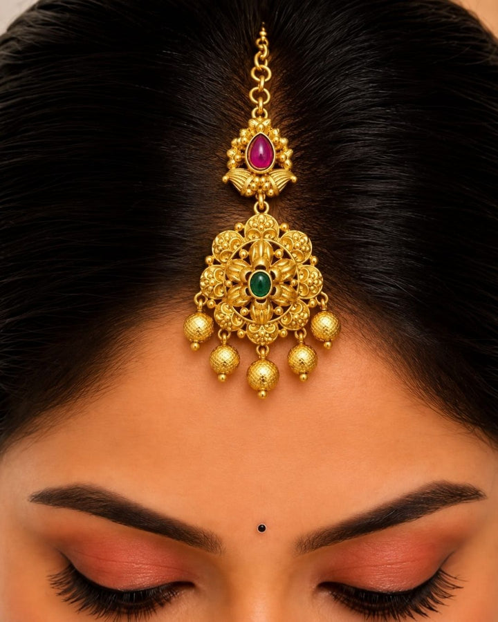 Traditional gold plated designer maangtikka featuring intricate floral motifs, green and ruby-colored stones, detailed gold work, and multiple hanging gold bead accents, perfect for festive, wedding, and traditional occasions.