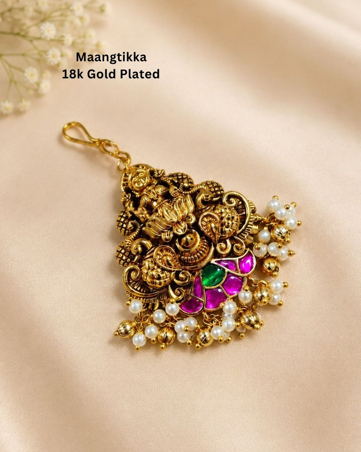 An ornate gold-plated Maangtikka hair pendant with intricate filigree work, small pearl-like beads, and pink and green stone accents, displayed on a beige satin background with text reading &