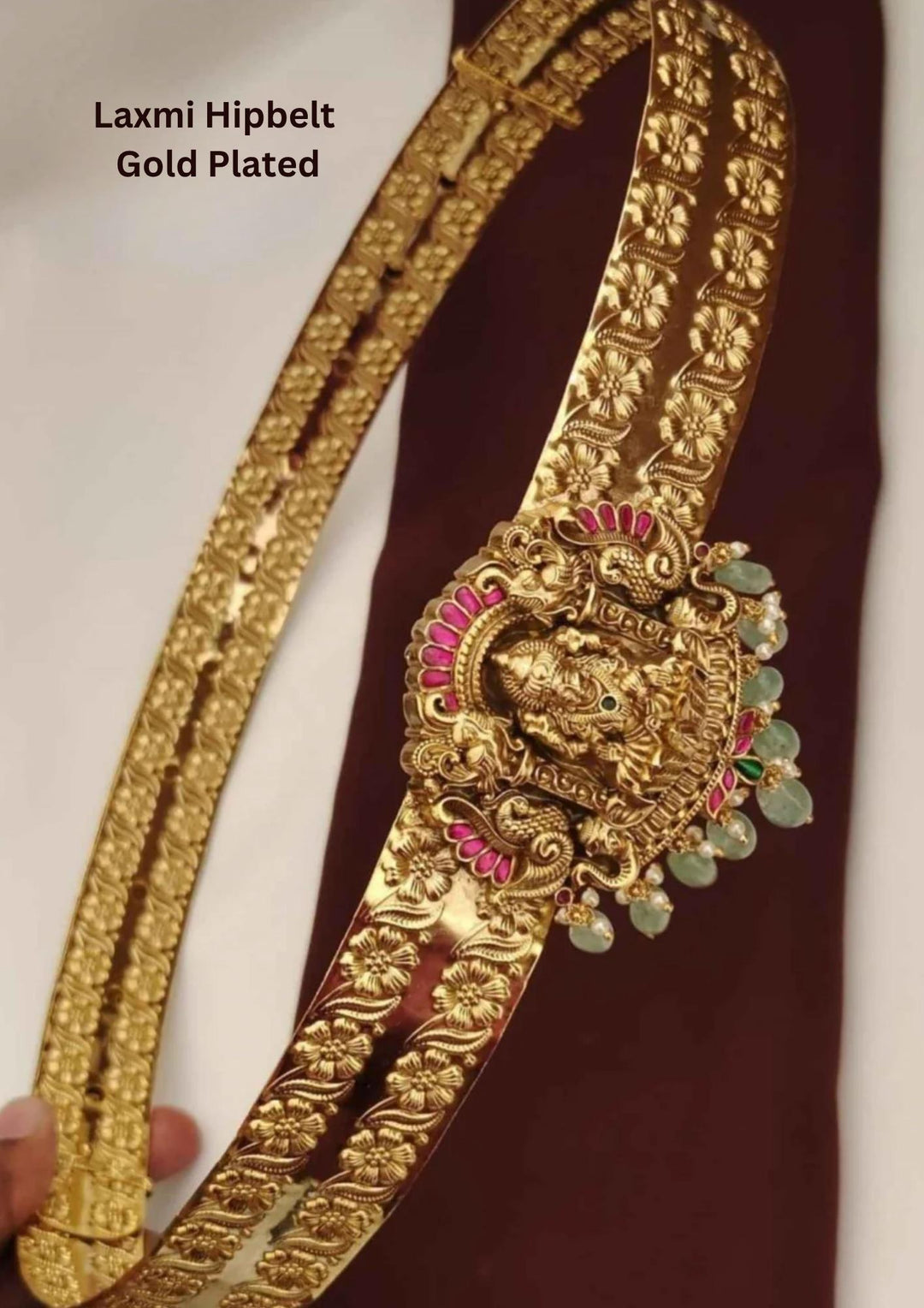 gold plated adult hipbelt vaddanam featuring two parallel flexible bands deeply embossed with repeating floral motifs. The central medallion showcases a detailed carving of Goddess Lakshmi flanked by twin peacocks inlaid with white crystals, emerald accents, and a fringe of hanging golden beads.