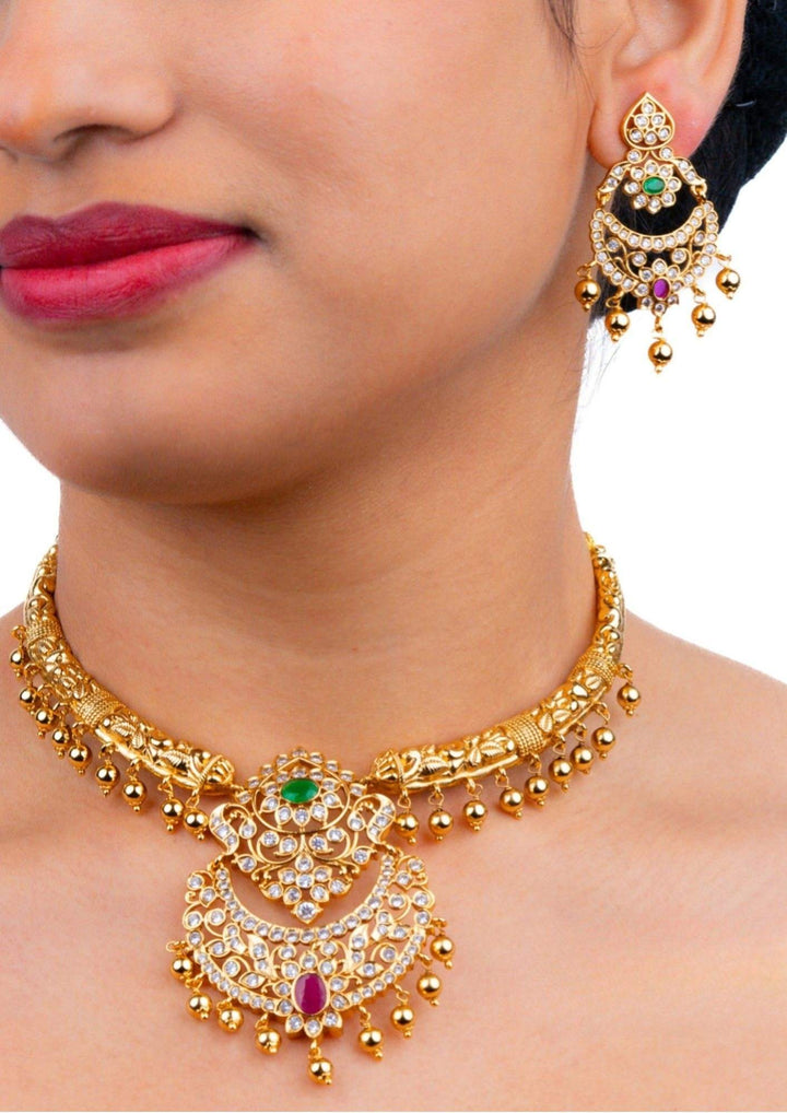 South indian women wearing Gold plated short antique necklace and matching hanging earrings set featuring structured metal blocks filled with clear white Jadau Kundan stones. The center of the necklace highlights a slightly larger floral design stone layout, and the entire lower rim of both pieces is decorated with neat, hanging clusters of small round golden-cream pearl beads, providing a vintage traditional look suitable for weddings and family functions.