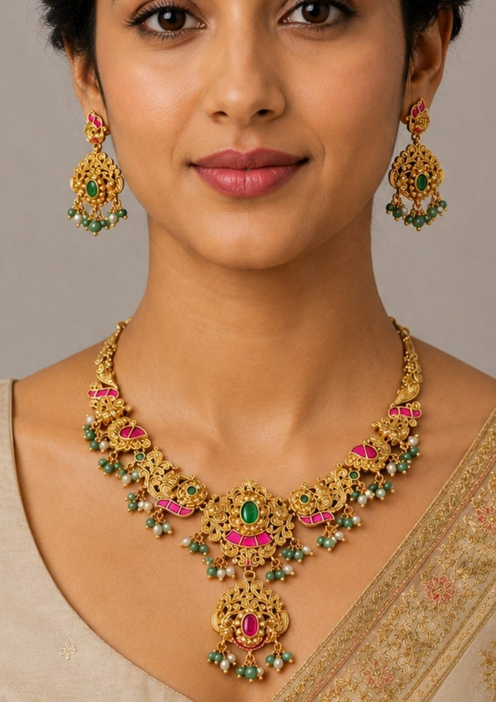 Woman wearing a gold necklace and earrings with green and red stones Traditional jadau kundan gold plated necklace set featuring intricate antique floral carvings with pink kundan accents, green center stones, pearl drops and green bead hangings. The heavy bridal necklace includes a grand pendant with matching chandbali style earrings, creating a rich South Indian traditional look suitable for weddings, bridal wear, temple functions and festive celebrations.