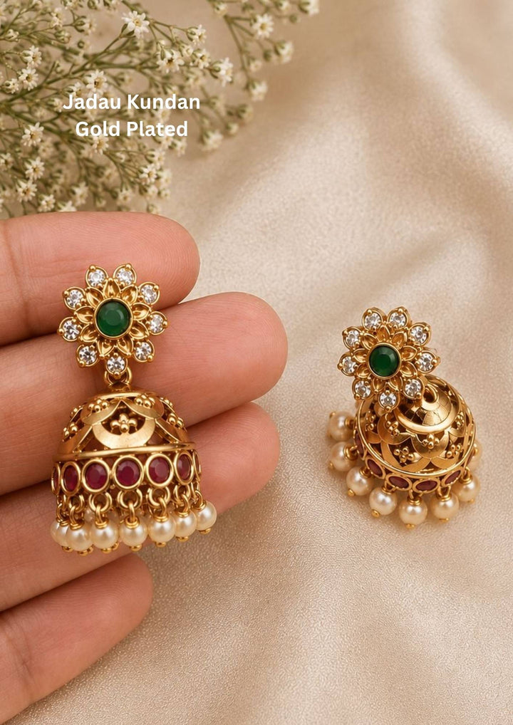 jadau kundan gold plated floral stud jhumka earrings featuring a top flower stud with an emerald green center surrounded by clear kundan stones, holding a beautifully carved gold bell dome lined with a bottom row of ruby red accents and delicate hanging white pearls. This lightweight classic jhumka pair is crafted with rich gold textures that provide a rich and classy appearance suitable for weddings, festivals, receptions, poojas, family functions
