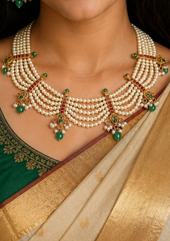 close up of  south indian neck waering a grand gold plated festive choker necklace meticulously hand-strung with five parallel tiers of smooth cream white rice pearls. The layered strands are joined by intricate vertical gold bars inlaid with red ruby squares and finish in dainty scalloped drapes accented by hanging green teardrop emerald beads and tiny pearl cluster bunches.