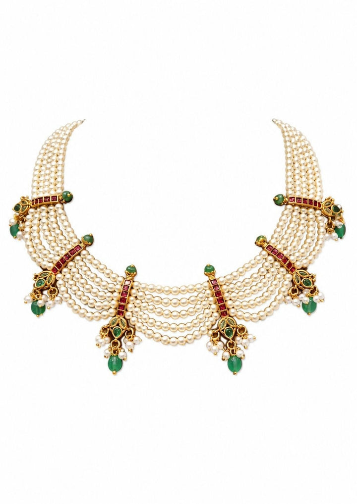 gold plated festive choker necklace meticulously hand-strung with five parallel tiers of smooth cream white rice pearls. The layered strands are joined by intricate vertical gold bars inlaid with red ruby squares and finish in dainty scalloped drapes accented by hanging green teardrop emerald beads and tiny pearl cluster bunches.
