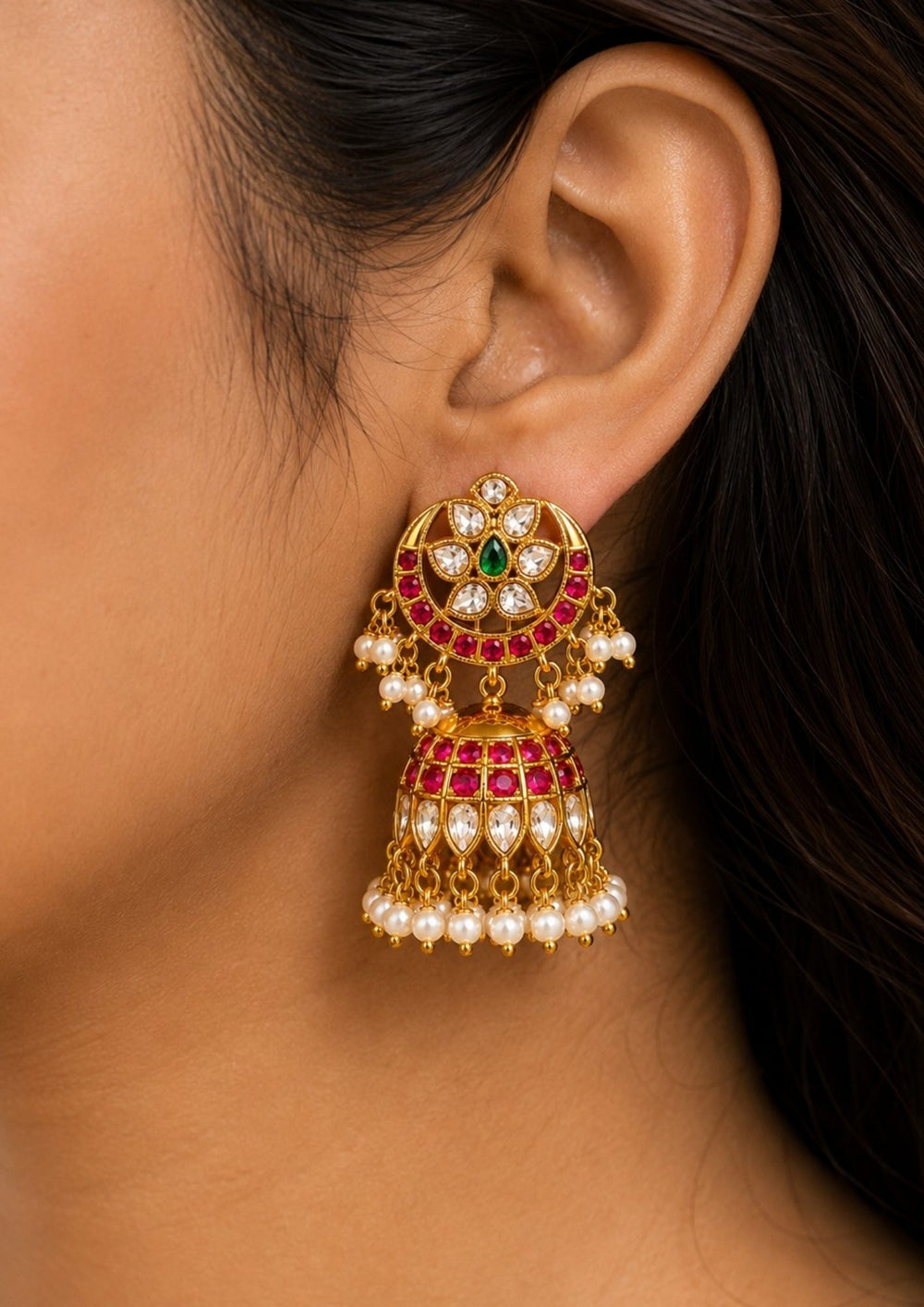 Gold earrings with red and green gemstones worn by model person Jadau Kundan gold plated jhumka earrings with detailed white kundan stones, ruby pink round stones, green center stone accents and hanging pearl drops in antique traditional design.  suitable for weddings, bridal wear, poojas, festivals, Bharatanatyam, temple functions, sarees and lehengas with lightweight comfortable fitting, anti fade polish, skin friendly quality