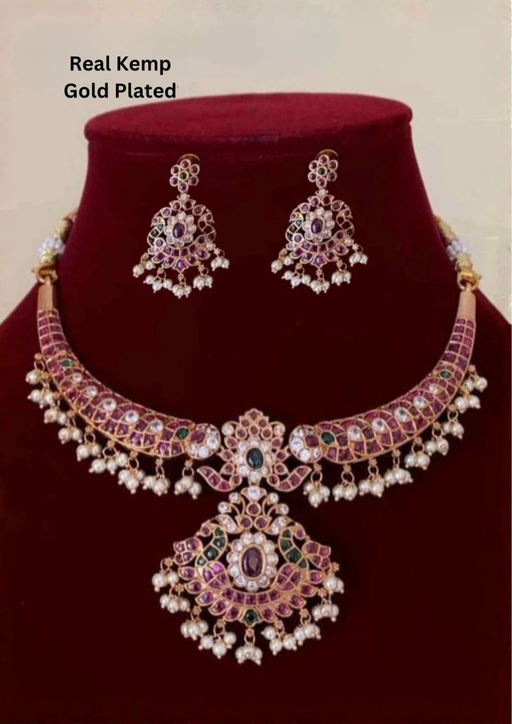 Jadau Kundan Gold Plated choker necklace and matching hanging earrings are designed with red ruby, emerald green, and white Kundan stones, finished with delicate white pearl guttapusallu upto the bottom borders. perfect for a rich, bridal-style look perfect for weddings, festivals, functions and grand look
