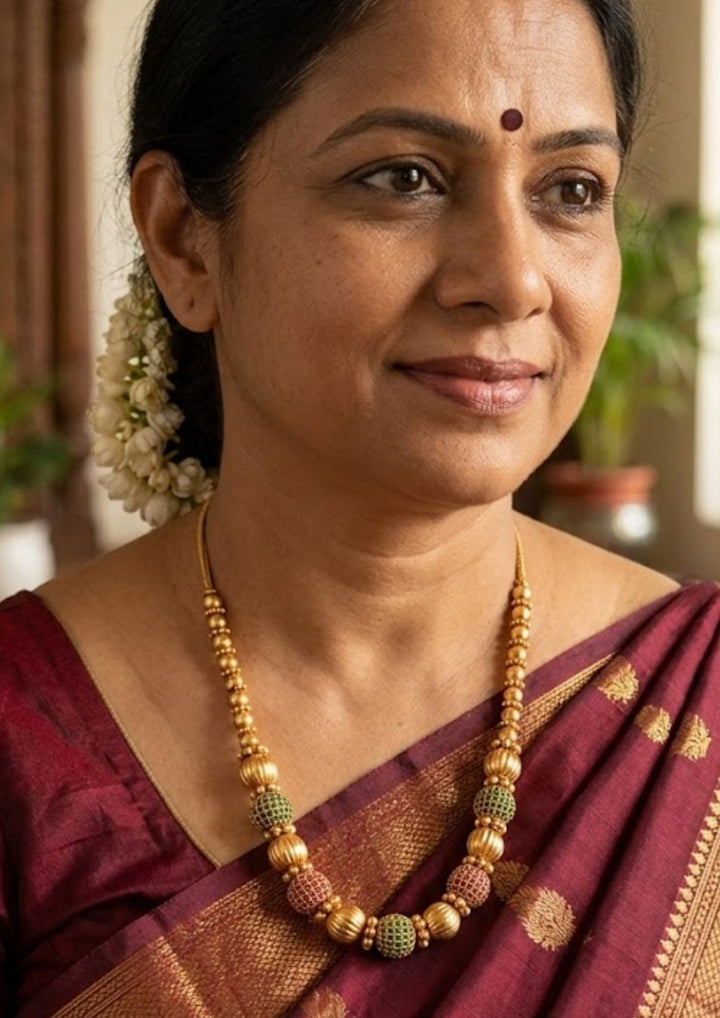 south indian women waering a Antique Gold Plated Beads Mala with long traditional chain designed with gold beads , antique finish pattern, and South Indian jewelry style. The mala comes with kundans and green and red stones in cube pattern and balls pattern with green stones . this is sweat proof quality, anti-fade colour, skin-friendly finish, and is suitable for pooja functions, festivals, daily wear, and traditional occasions. 