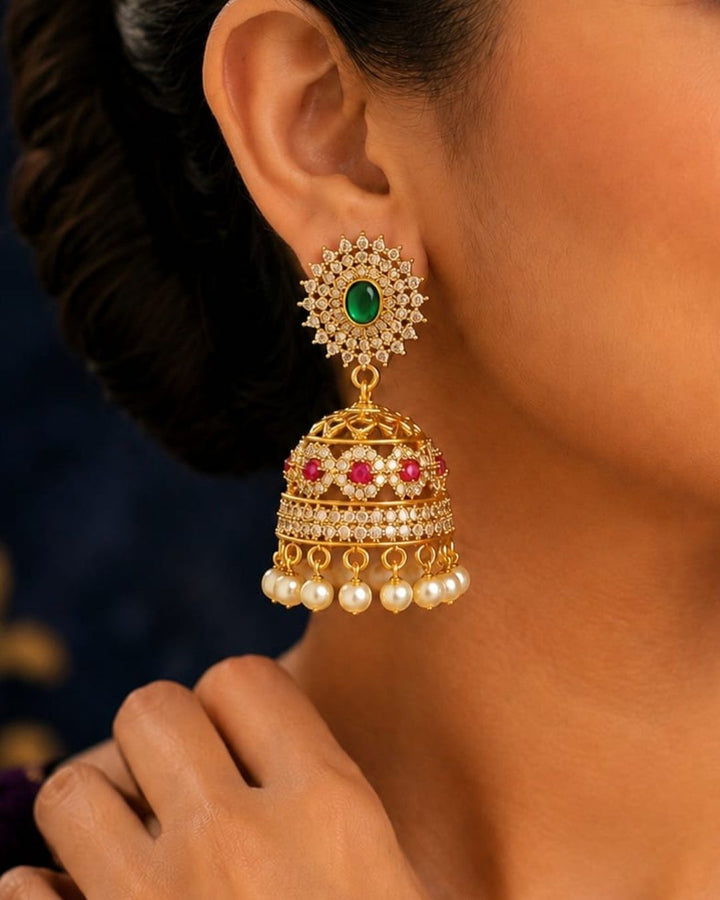 A close-up of a woman wearing a traditional Indian gold jhumka earring with a green stone, red stones, and white pearls.