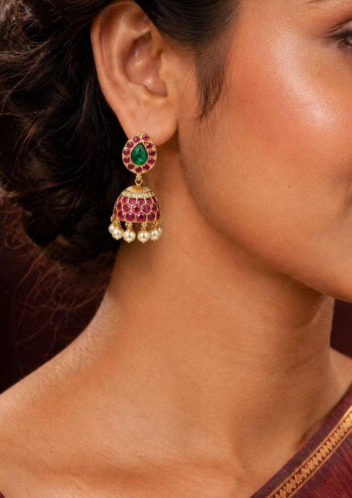 Close-up of a south indian women wearing a Exquisite Jadau Kundan gold-plated jhumka earrings featuring a paisley or teardrop-shaped stud top. The top is centered with a striking green emerald stone framed by vibrant ruby-pink stones, leading down to a honeycomb-patterned dome adorned with pink stones and finished with elegant hanging pearl drops. Perfect for weddings and festive celebrations.