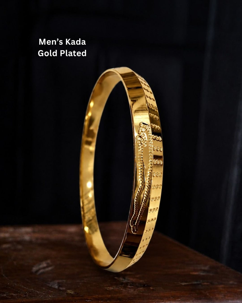 A gold-plated men's kada bangle bracelet with engraved patterns, displayed on a dark wooden surface against a black background with text overlay reading 'Men's Kada Gold Plated'.