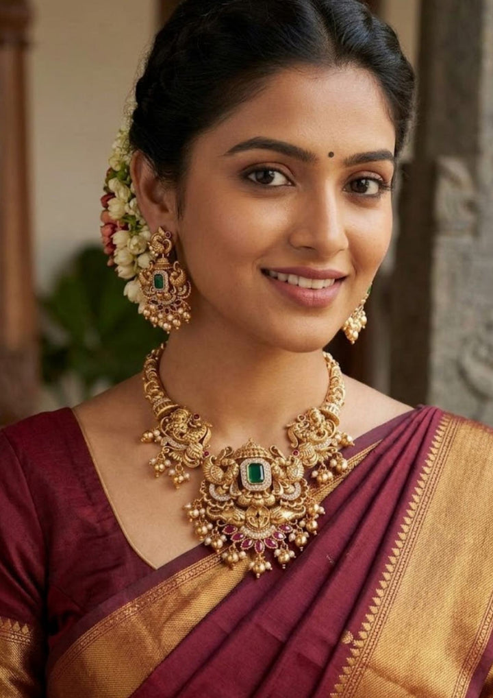Close-up of a south indian women wearing a premium 18K gold plated Kundan Emerald antique temple necklace and matching grand earrings beautifully detailed with Lakshmi motifs, ruby pink accents, and dangling pearl clusters for a classic bridal look.