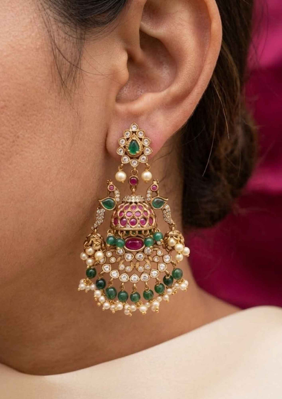 Close-up of south indian woman earring with Jadau Kundan gold plated heavy bridal earrings.The design features a teardrop top stud with a green stone center, connecting to dual peacock motifs flanking a main dome-shaped jhumka. Below the dome is a wide, semi-circular crescent layout encrusted with white kundan stones, layered with hanging dark green beads and guttapusallu. perfect for weddingwear, festivals and it is anti fade color