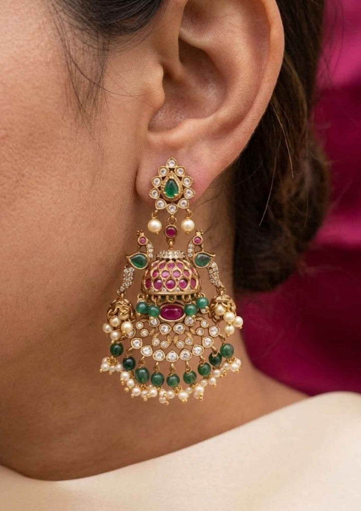 Close-up of south indian woman earring with Jadau Kundan gold plated heavy bridal earrings.The design features a teardrop top stud with a green stone center, connecting to dual peacock motifs flanking a main dome-shaped jhumka. Below the dome is a wide, semi-circular crescent layout encrusted with white kundan stones, layered with hanging dark green beads and guttapusallu. perfect for weddingwear, festivals and it is anti fade color