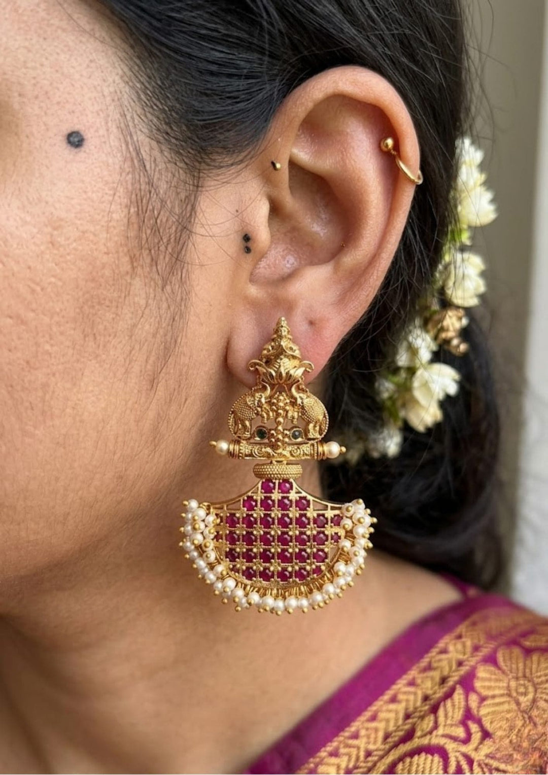 South indian women waering Gold plated hanging earrings featuring a structured top section beautifully embedded with clear, shimmering white Kundan stones. The center highlights vibrant red or green kemp stones structured in a classic floral or crescent frame, while the lower section finishes with an elegant, swinging bunch of real smooth gemstone beads, providing a colorful heritage look perfect for weddings and festivals.
