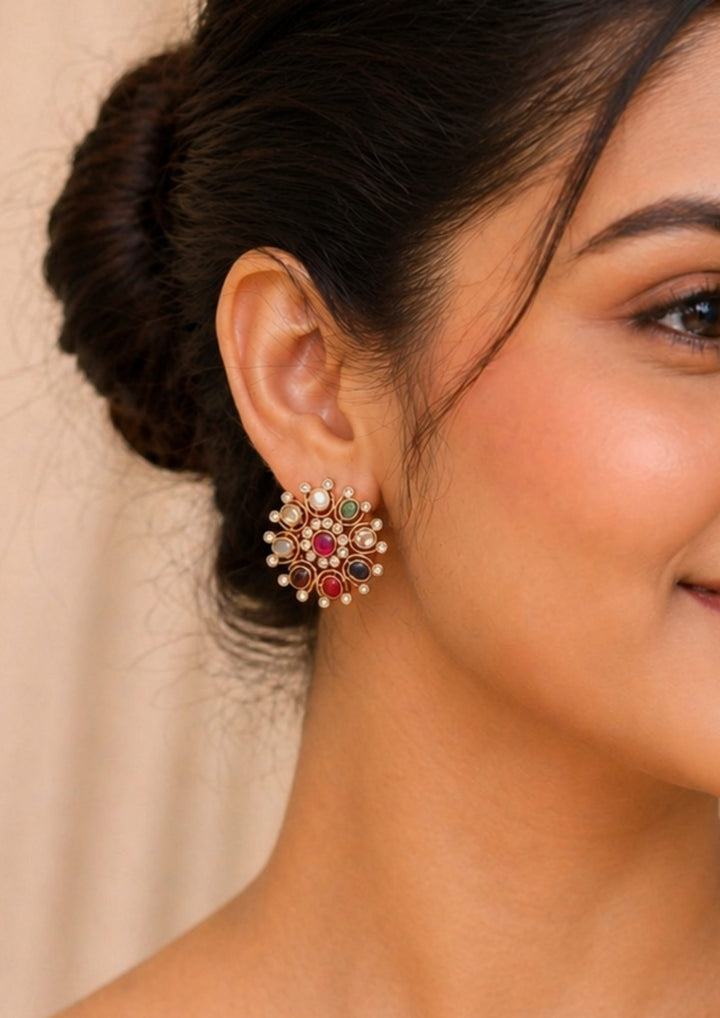 Close-up of a south indian woman wearing a Traditional Navaratna gold plated big stud earrings featuring a classic circular flower design stamped with nine auspicious multi-colored stones. The center features a prominent pink ruby gemstone surrounded by a delicate halo of small white CZ crystals and various colored oval gems—including blue, green, yellow, red, and pearl white—each tipped with a tiny shimmering white stone.