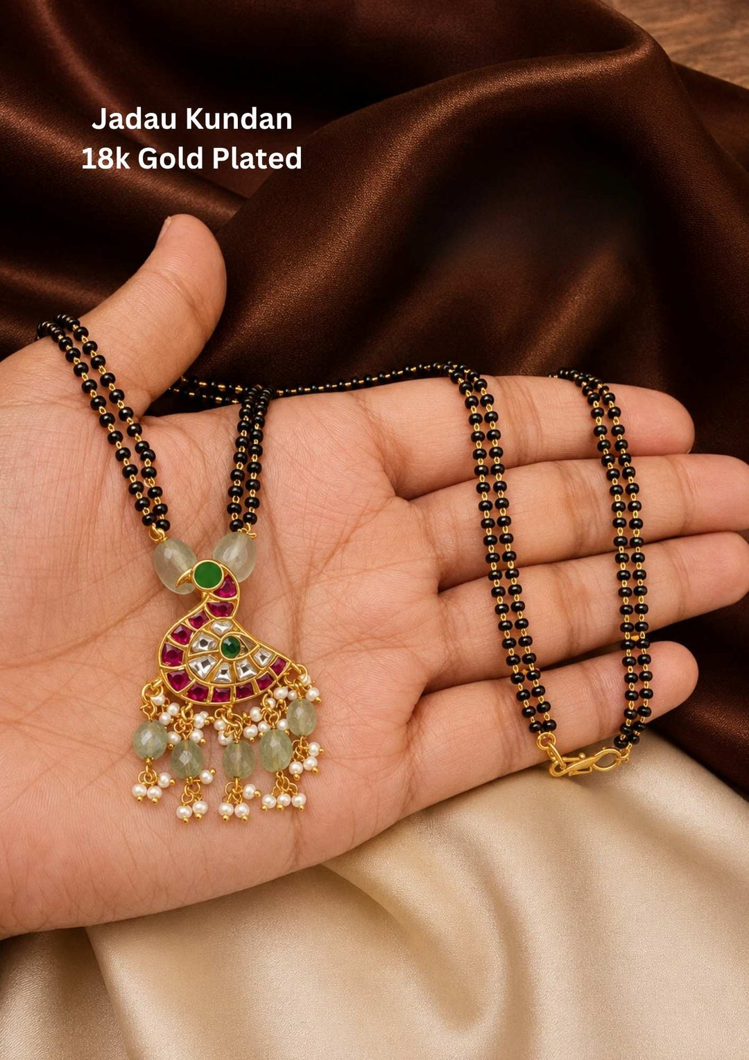 gold plated Jadau Kundan mangalsutra featuring a classic double-strand black beaded chain. The chain holds an artistic peacock-shaped pendant inlaid with white kundan crystals, ruby-red borders, and green enamel accents. The base of the pendant is finished with a delicate fringe of light green tumble beads and tiny white pearl clusters. Designed for daily or festive wear with premium quality, comfortable fitting, and Cash on Delivery available.