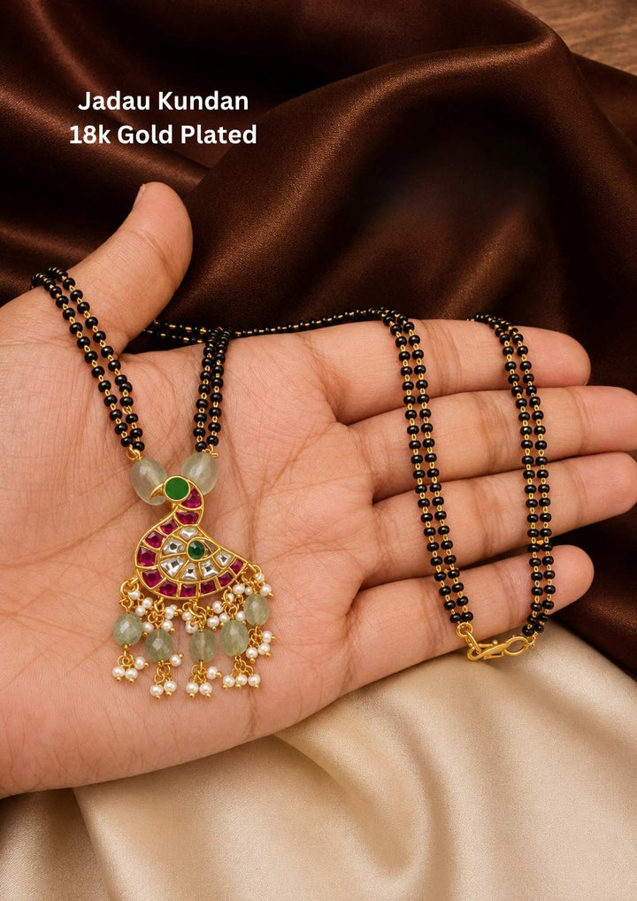 gold plated Jadau Kundan mangalsutra featuring a classic double-strand black beaded chain. The chain holds an artistic peacock-shaped pendant inlaid with white kundan crystals, ruby-red borders, and green enamel accents. The base of the pendant is finished with a delicate fringe of light green tumble beads and tiny white pearl clusters. Designed for daily or festive wear with premium quality, comfortable fitting, and Cash on Delivery available.