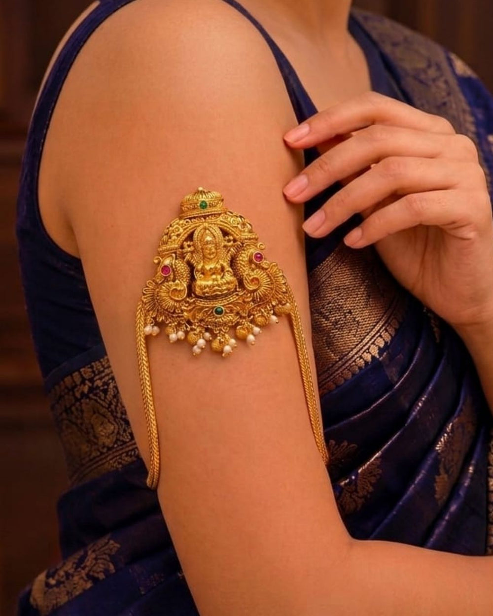 Traditional gold plated Vanki armlet featuring an ornate Goddess Lakshmi design, intricate floral detailing, green and red stone accents, and delicate pearl and gold bead hangings.